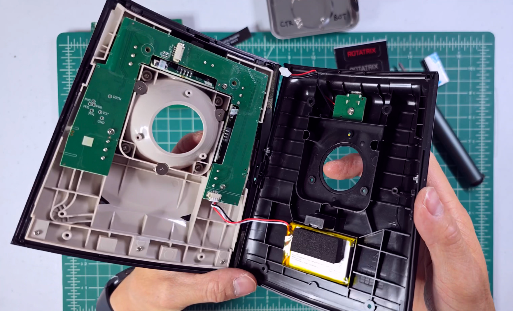 The opened case with the two shell halves apart, showing the battery and the two wire-to-board connectors to disconnect from the board